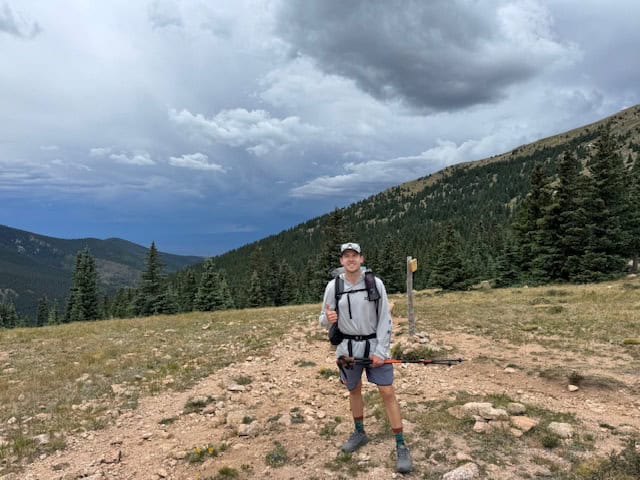 Hiker standing on trail in mountainous landscape with cloudy sky.